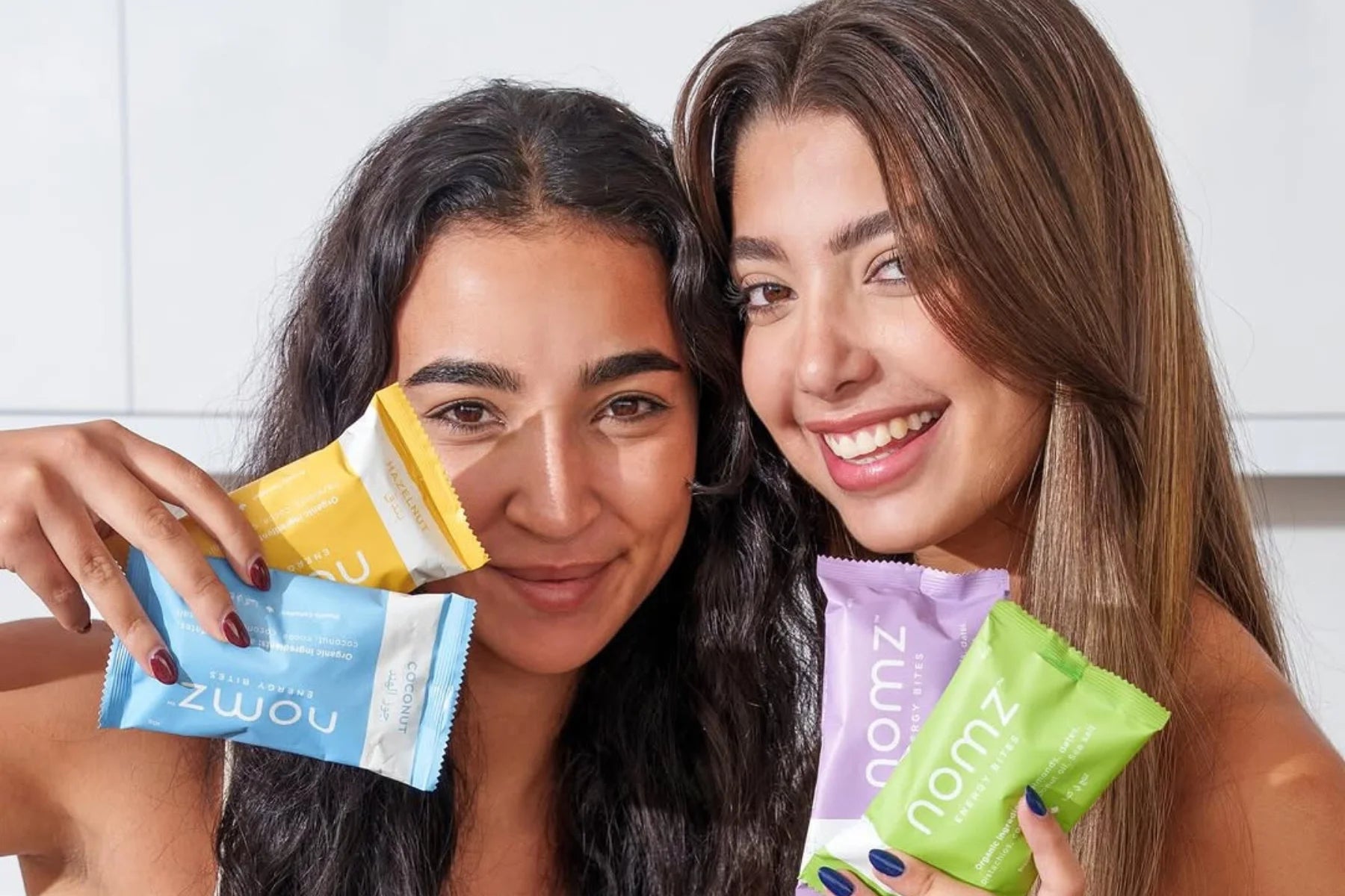Two models showcasing the four NOMZ variant packs (Almond, Pistachio, Hazelnut, and Coconut).